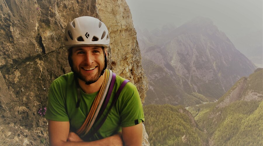 Kletterer mit Helm lehnt an einer Felswand mit Blick auf bewaldete Berge im Hintergrund. | © Baumgartner Martin
