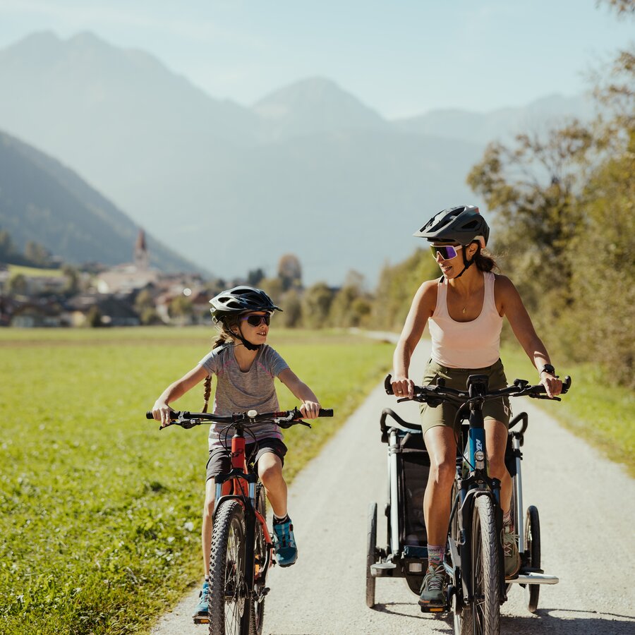 Mother and child ride side by side with a bike trailer, village of Oberrasen in the background. | © IDM Südtirol - Alex Molling