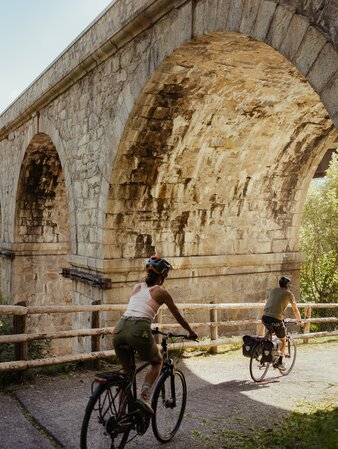 Due ciclisti percorrono un sentiero sotto un viadotto in pietra. | © IDM Südtirol - Alex Molling