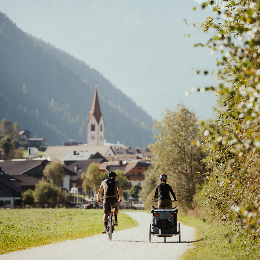Two cyclists with a child trailer ride toward the village of Oberrasen with church and mountains in the background. | © IDM Südtirol - Alex Molling
