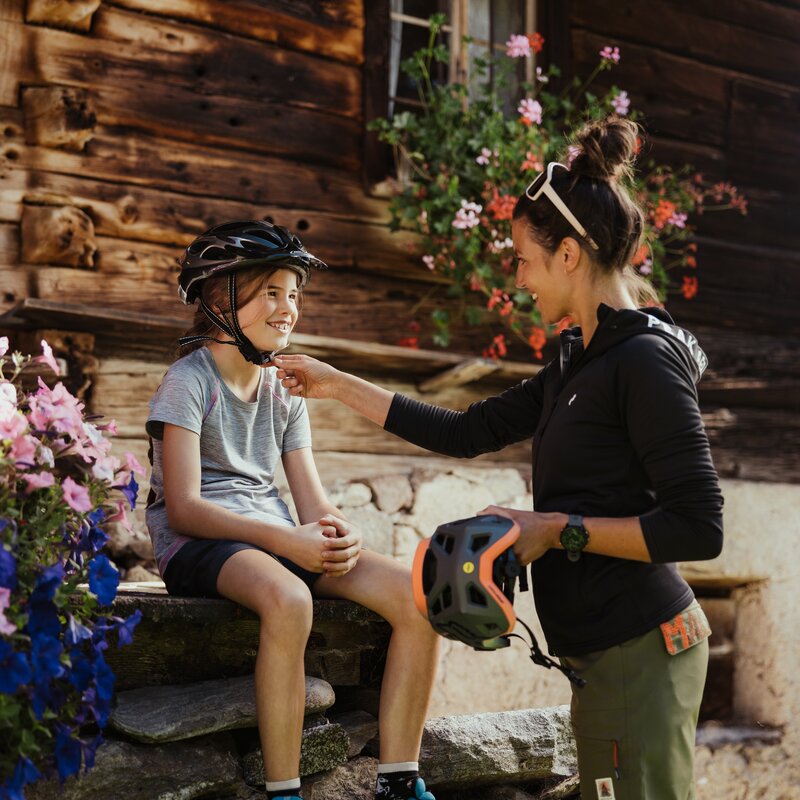 Eine Frau hilft einem Kind beim Anlegen des Fahrradhelms vor einem alten Bauernhaus mit Blumen. | © IDM Südtirol - Alex Molling